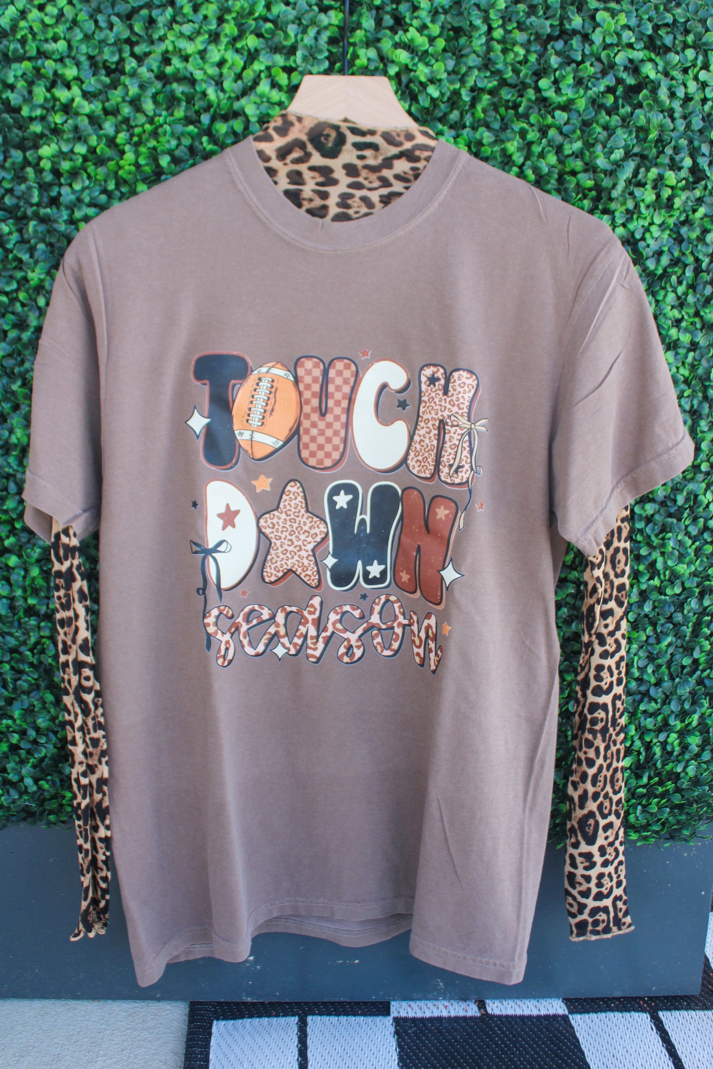 Brown t-shirt with colorful text and patterns on a mannequin against a green leafy background