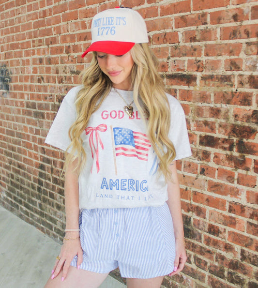 Woman wearing a graphic t-shirt and cap with text and an American flag design, standing against a brick wall.