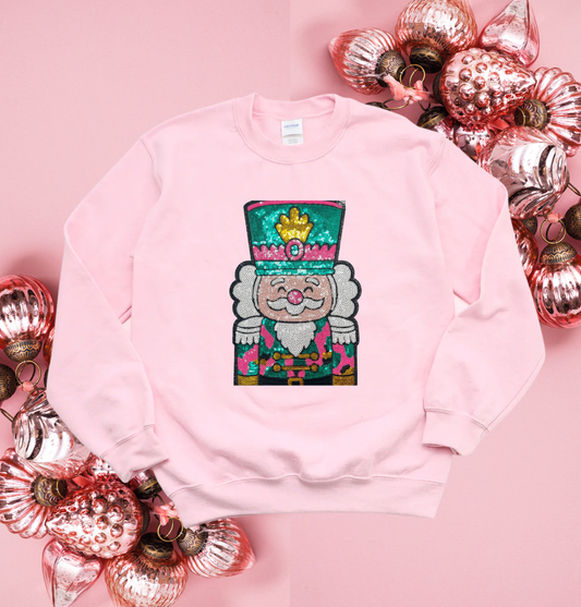 Pink sweatshirt with a colorful nutcracker design on a pink background with decorative ornaments.