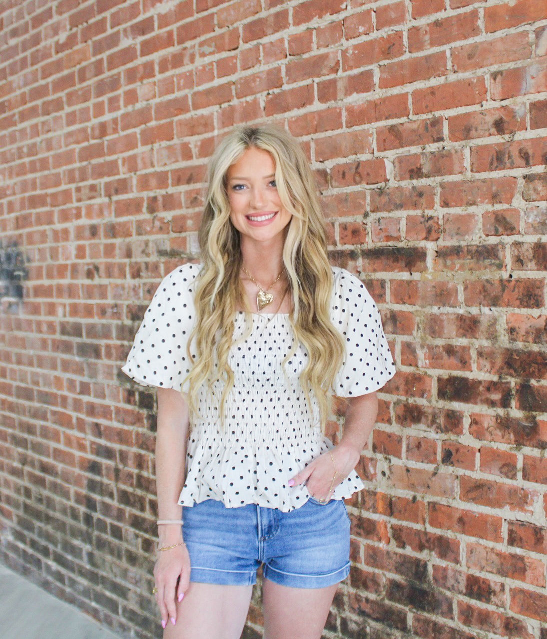 Woman wearing a polka dot top and denim shorts standing against a brick wall.