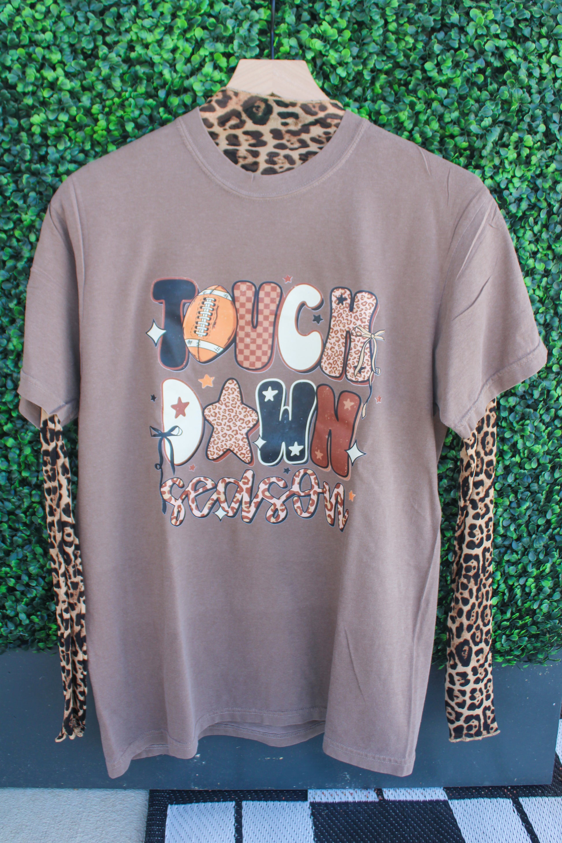 Brown t-shirt with colorful text and patterns on a mannequin against a green leafy background